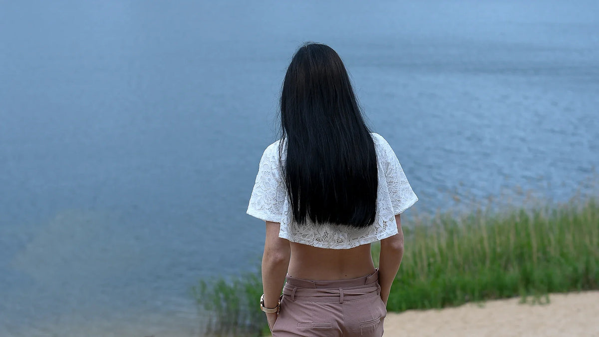 A woman in a white top and pink trousers standing beside calm water, seen from behind.