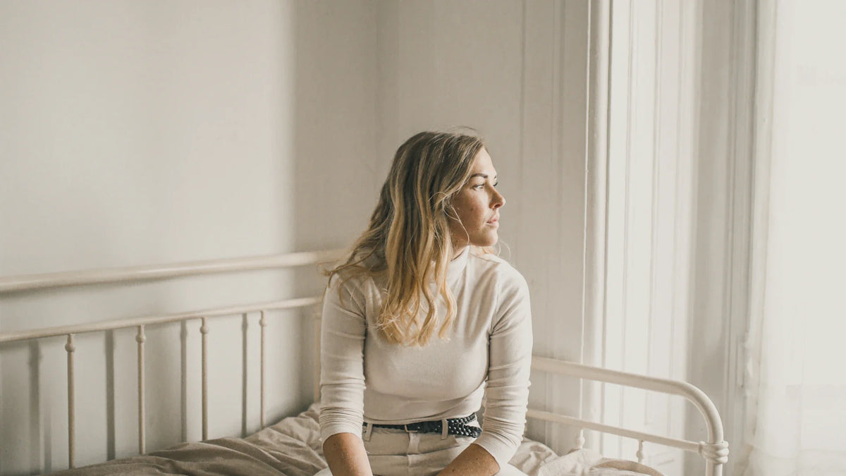A woman sits quietly on a bed near a bright window.
