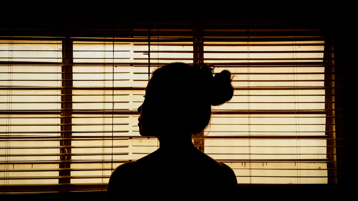 An adult woman’s dark silhouette beside horizontal window blinds.