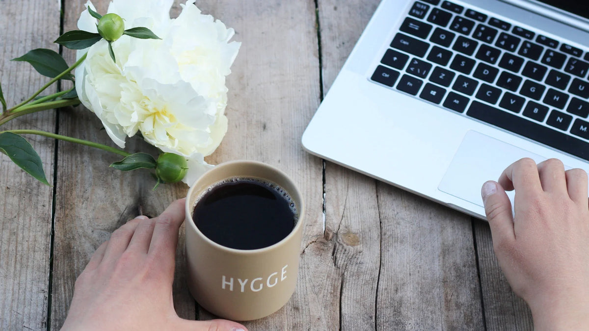 Hands beside a coffee cup, laptop and pale flower on a table.