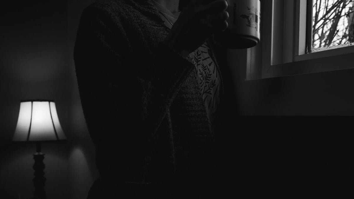 A shadowed figure holding a mug beside a window, with no face visible in the crop.