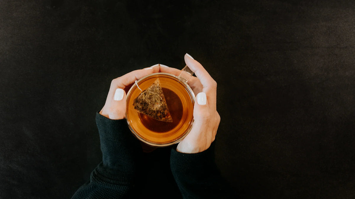 Cropped hands holding a clear glass cup of tea against a dark background.