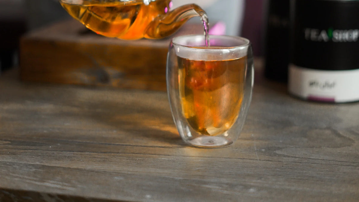 Tea being poured from a glass teapot into a double-walled cup on a wooden table.