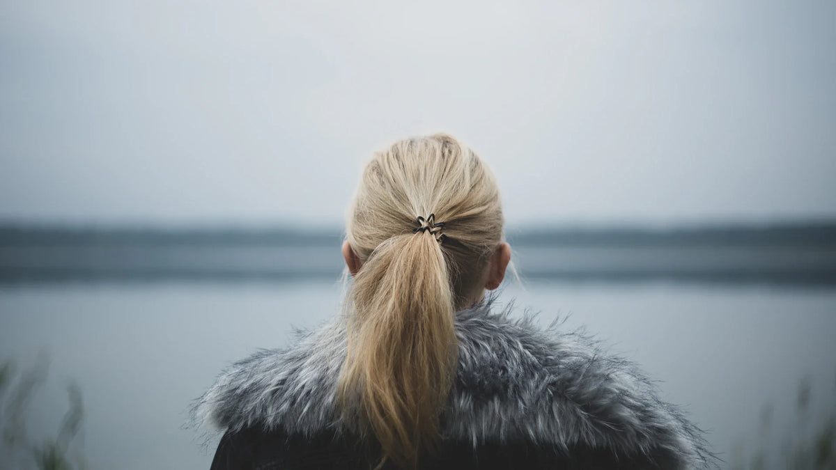 An adult woman viewed from behind beside a lake in soft grey light.
