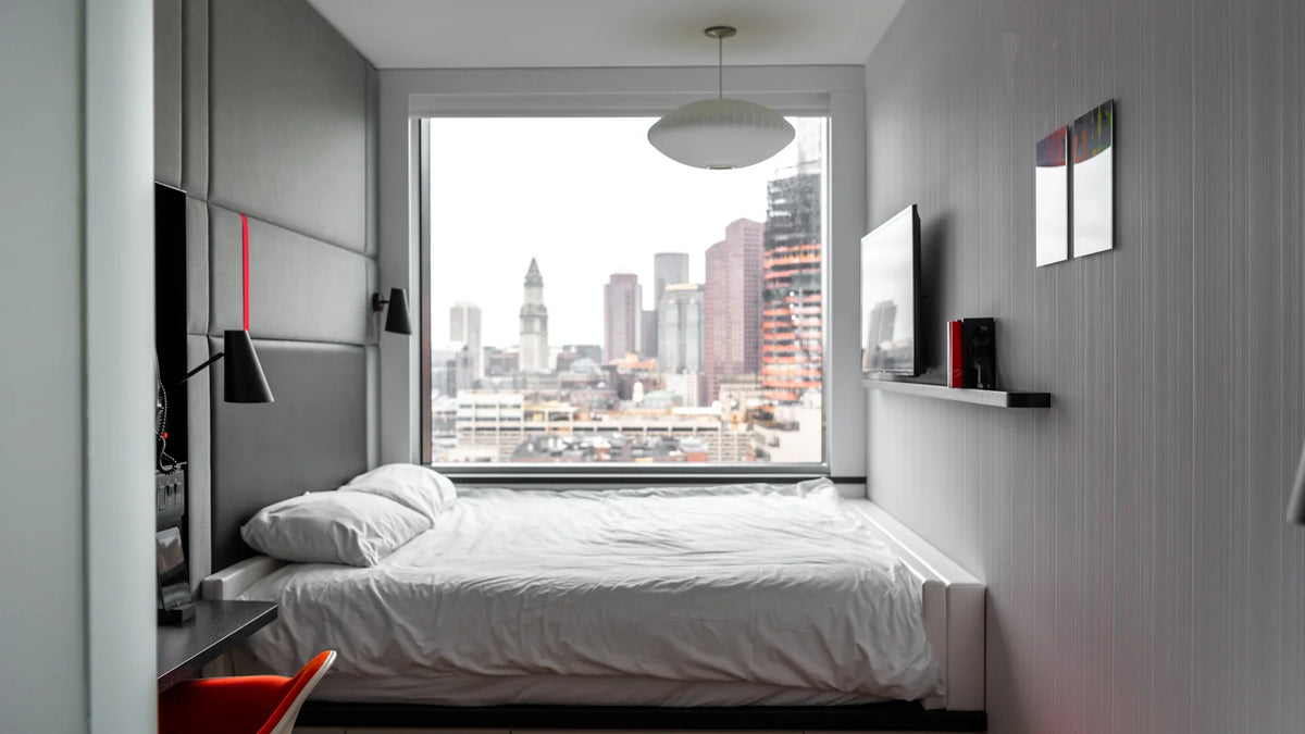 White bedding beside a large window overlooking a city.