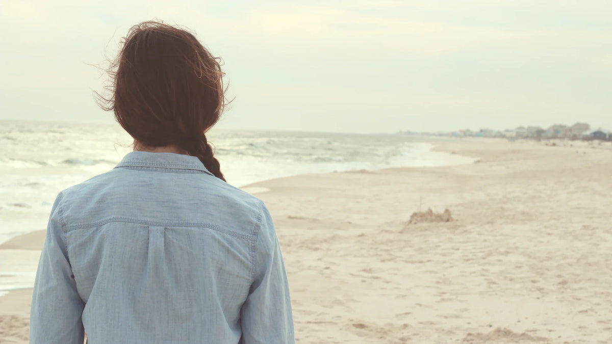 A woman in a pale blue shirt looking out to sea, photographed from behind.