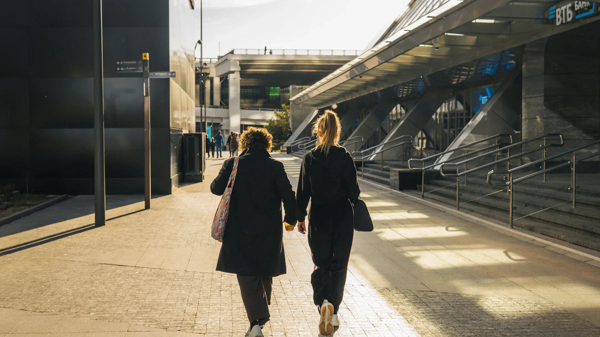 Two adult women walking together through a sunlit city, seen from behind.
