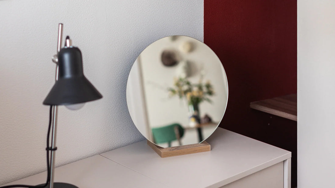 A bedside table with a lamp and a round mirror in a bedroom.