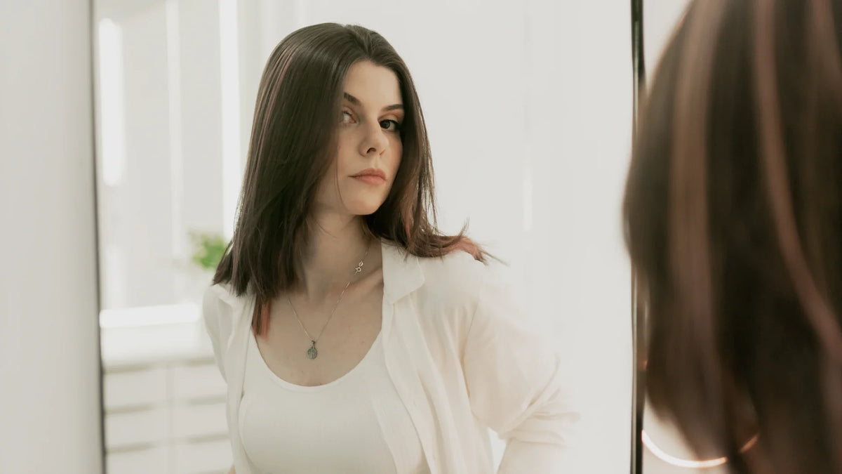A fully clothed woman stands by a mirror in a bright room.