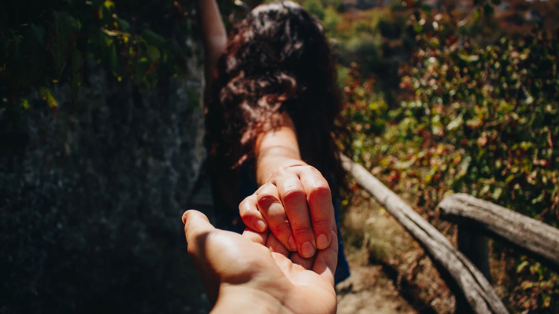 A woman holds another person’s hand outdoors, seen from behind.