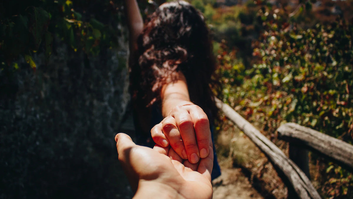 A woman holds another person’s hand outdoors, seen from behind.