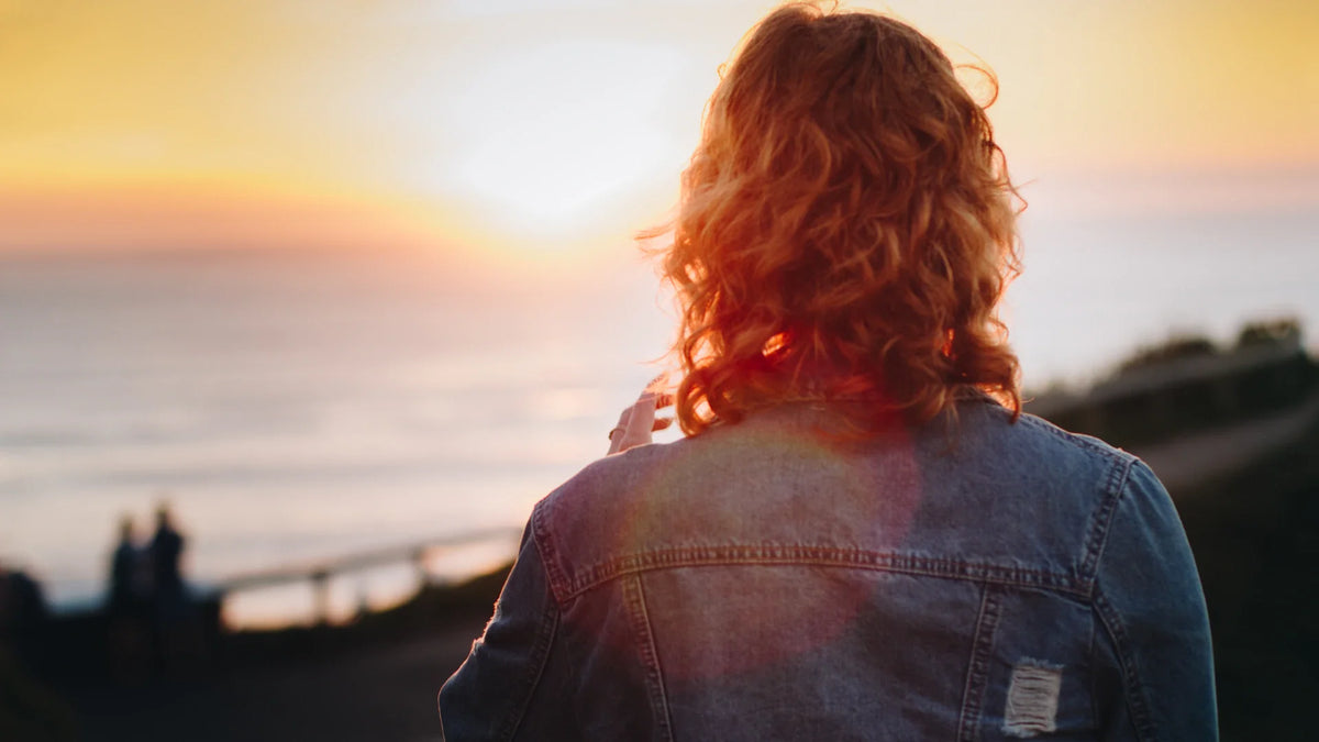 A woman in a denim jacket looks toward the sea at sunset, photographed from behind.