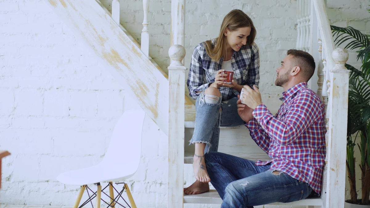 Two adults sit on stairs at home and talk over warm drinks.