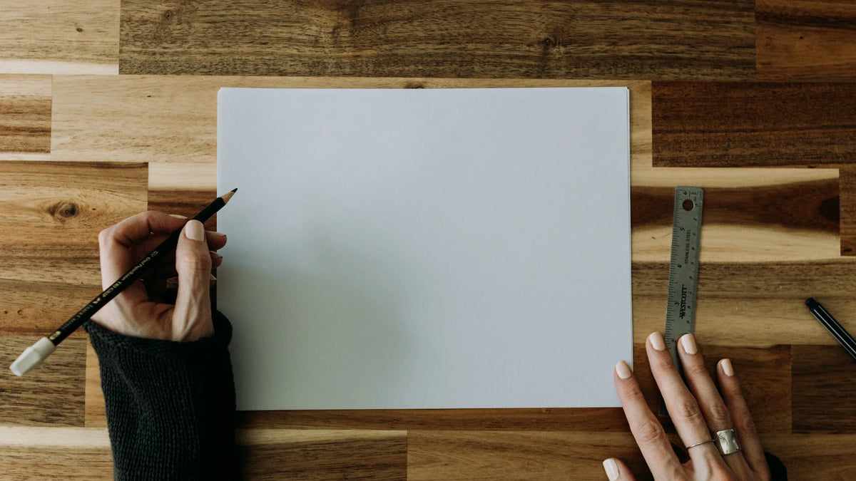 Cropped hands holding a pencil beside blank paper and a ruler on a wooden table.