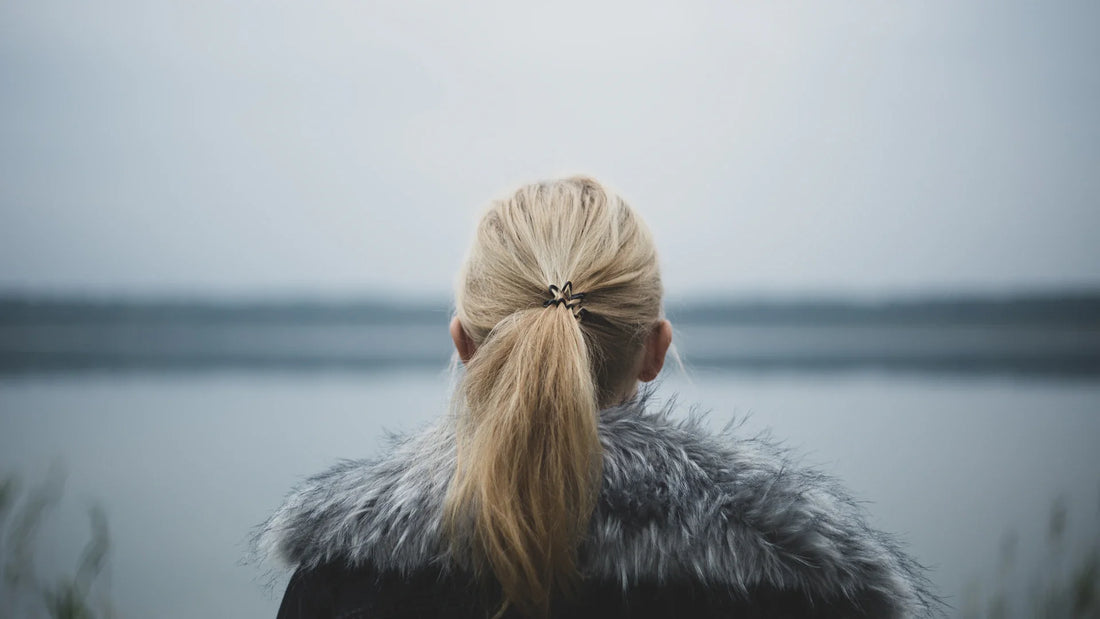 An adult woman viewed from behind beside a lake in soft grey light.