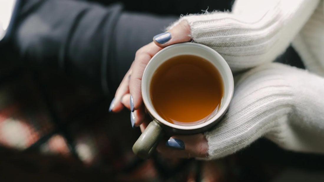 Cropped hands with purple nails holding a cup of tea beside a white sweater.