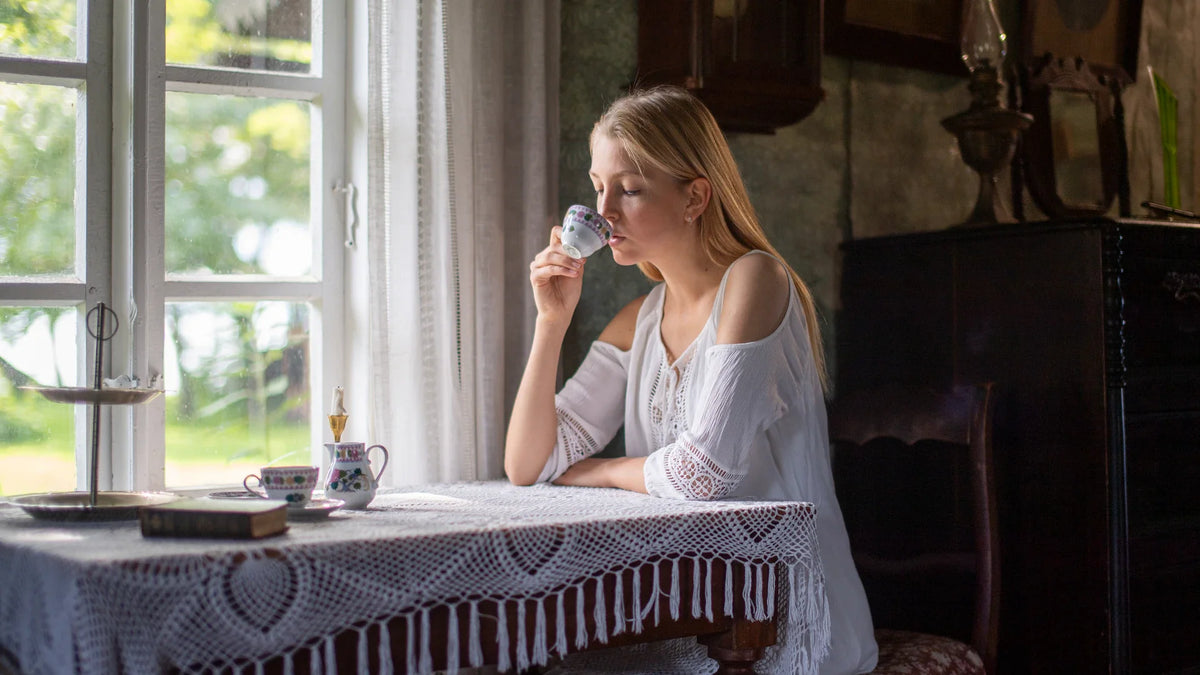 A woman drinks tea beside a window.