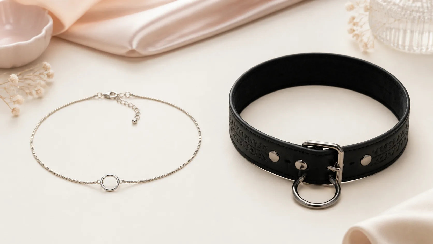 Slim silver jewelry-like day collar beside a wider unfastened black adult play collar on a warm neutral surface