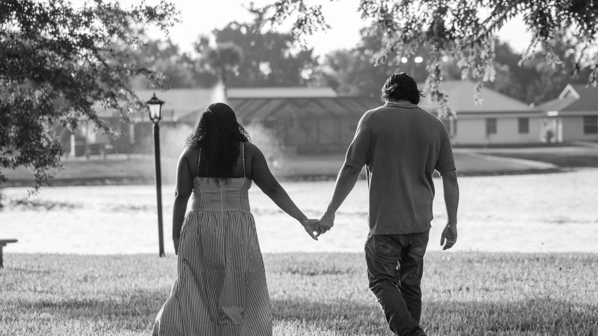 A couple walking hand in hand beside a lake, photographed from behind in black and white.
